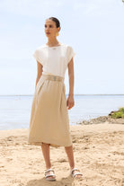 Woman wearing a midi dress with a fluid skirt and cap sleeves, standing on a sandy beach with water and sky in the background.
