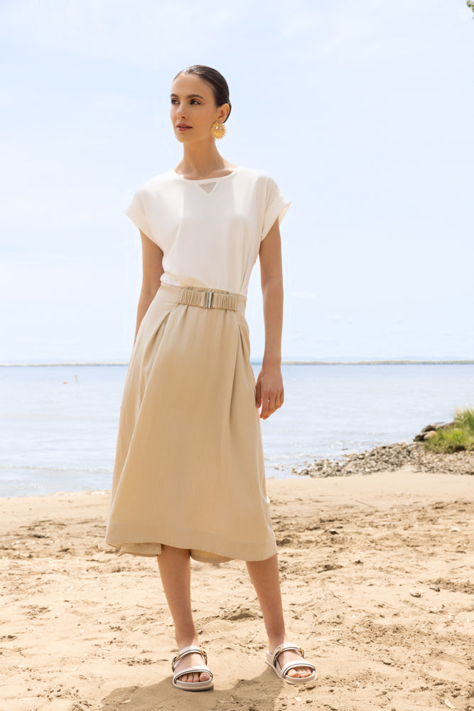 Woman wearing a midi dress with a fluid skirt and cap sleeves, standing on a sandy beach with water and sky in the background.