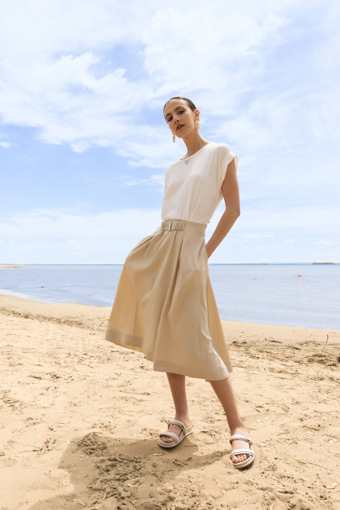 Woman wearing a midi dress with a fluid skirt and cap sleeves, standing on a sandy beach with water and sky in the background.