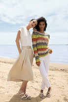 Two women standing on a beach near the water; one wears a midi dress with a relaxed fit and belted waist, while the other wears a textured long-sleeve sweater with slim pants and flat sandals.