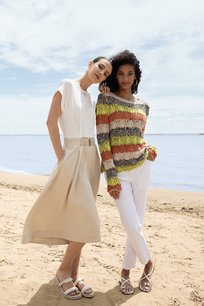 Two women standing on a beach near the water; one wears a midi dress with a relaxed fit and belted waist, while the other wears a textured long-sleeve sweater with slim pants and flat sandals.