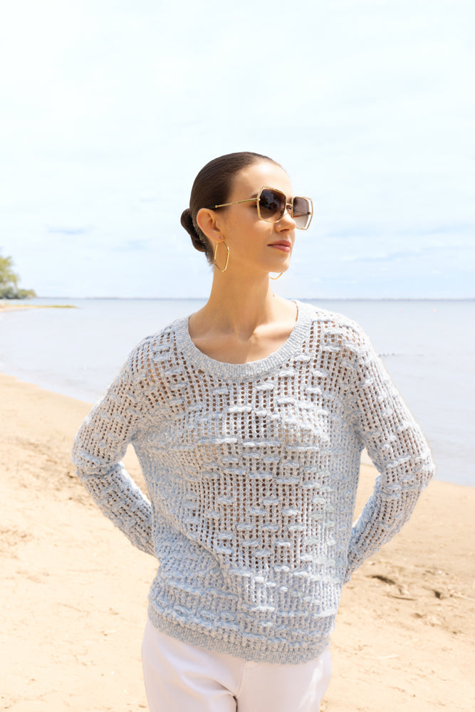 Model wearing a light blue open-knit crewneck sweater with long sleeves and ribbed trim, styled with light trousers and sunglasses at the beach.