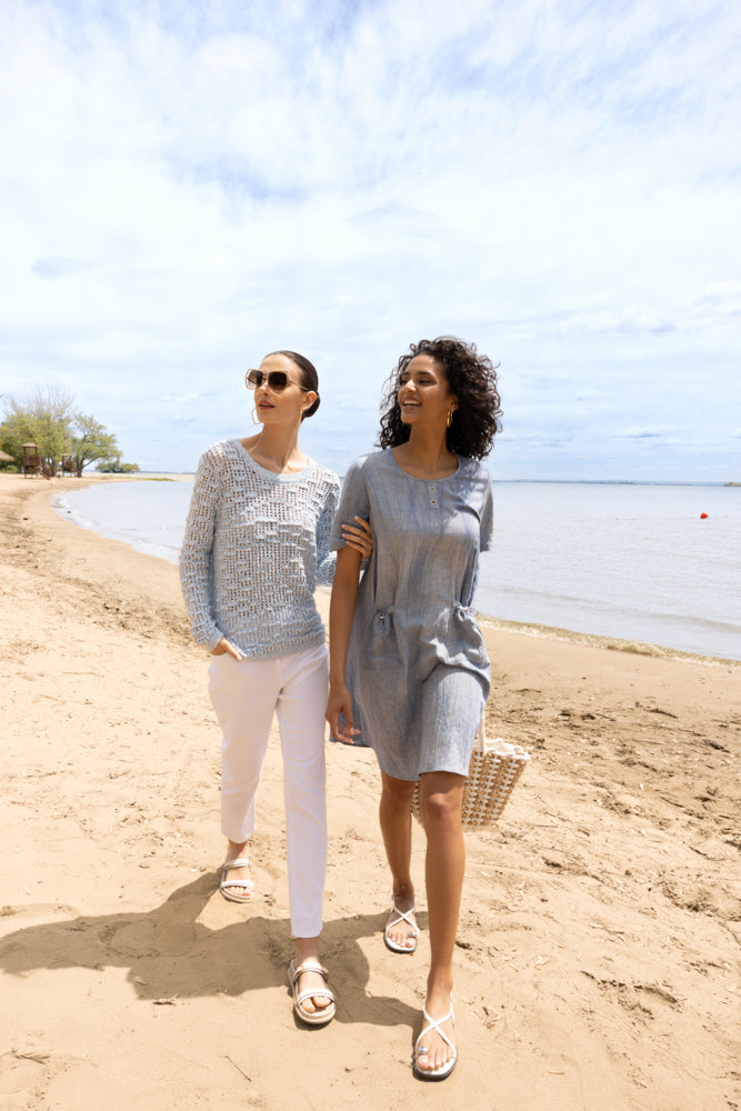 Two women walking along a sandy beach wearing casual summer outfits—one in a light blue open-knit sweater with white pants and beige sandals, the other in a grey short-sleeve dress with white sandals and carrying a woven tote bag.