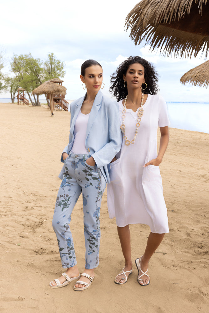 Model wearing a light blue linen-blend blazer with matching floral slim-leg trousers and a white top, styled with neutral sandals on a sandy beach.