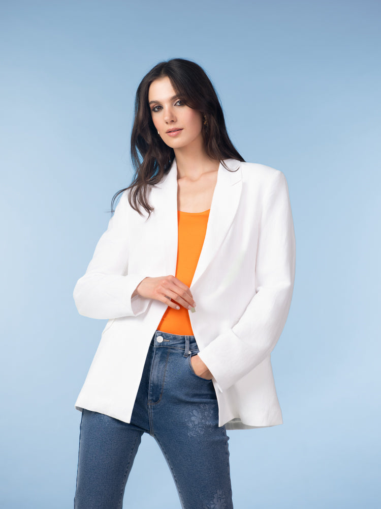 Model wearing a white linen-blend blazer layered over a bright orange sleeveless top and fitted blue jeans against a studio backdrop.