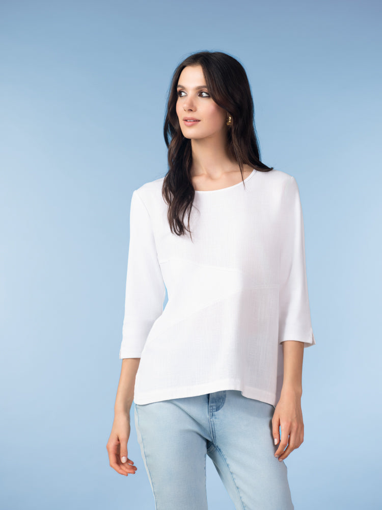 Model wearing a white three-quarter sleeve blouse with angled panel detailing, paired with light wash jeans, front-facing against a soft blue studio background.