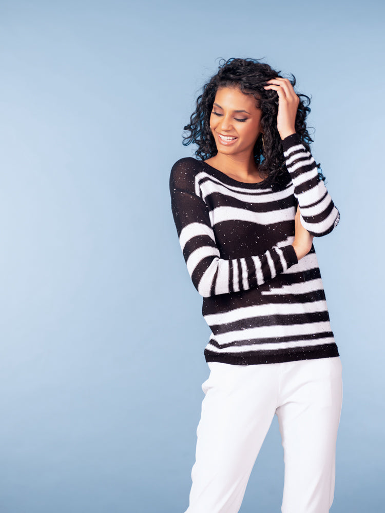 Model wearing a black and white striped lightweight knit sweater with subtle shimmer, styled with white trousers against a light blue background.
