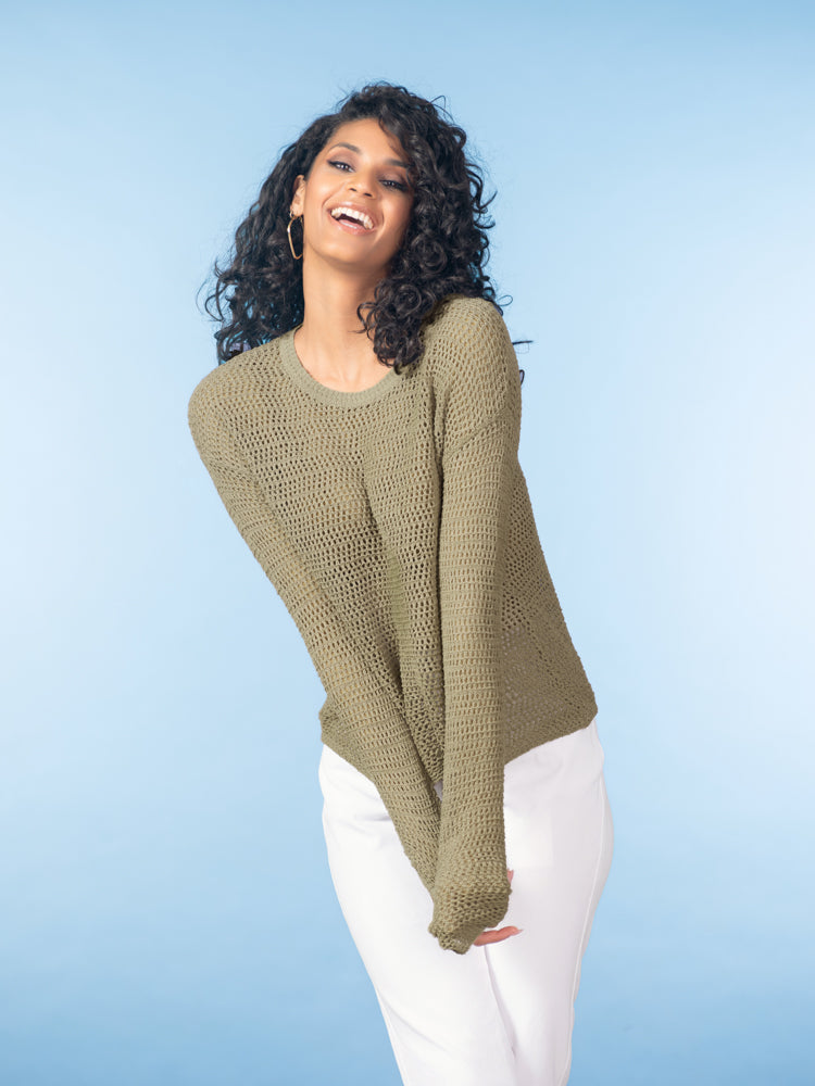 Model wearing a khaki open-knit long-sleeve sweater styled with white trousers against a soft blue studio background.
