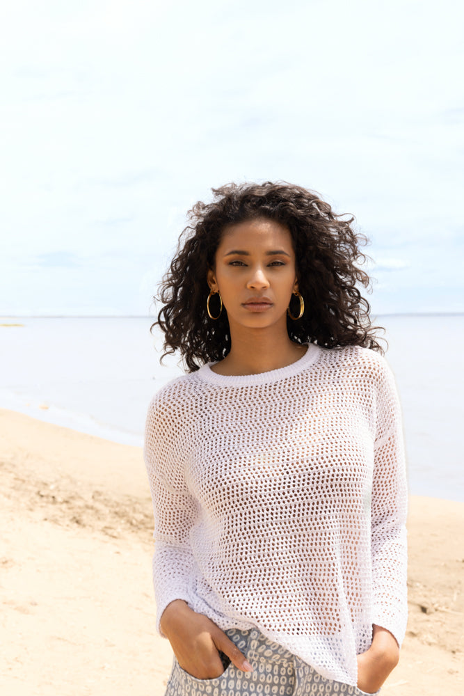 Model posing on a beach wearing a white open-knit long-sleeve sweater paired with a light blue patterned knee-length skirt and white sandals.
