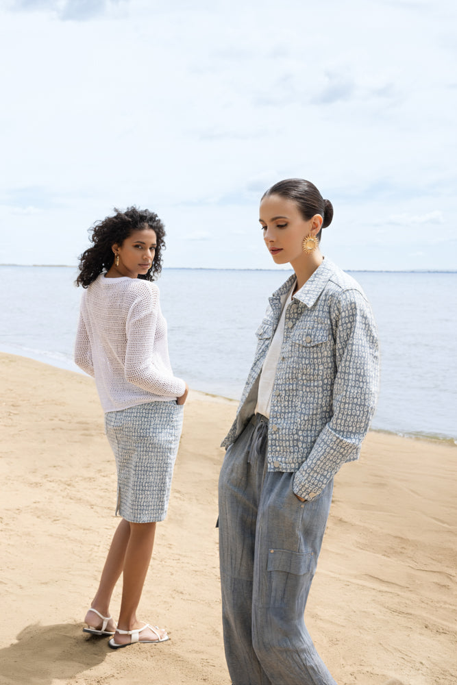 Two models standing on a sandy beach wearing lightweight open-knit sweaters; one styled with a white mesh sweater and printed knee-length skirt, the other in a patterned jacket with wide-leg trousers.