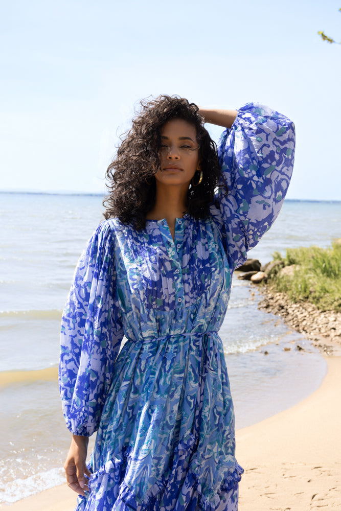 Close-up of a woman in a blue and white abstract floral midi dress with button-front neckline and waist tie, standing by the water with natural shoreline in the background.