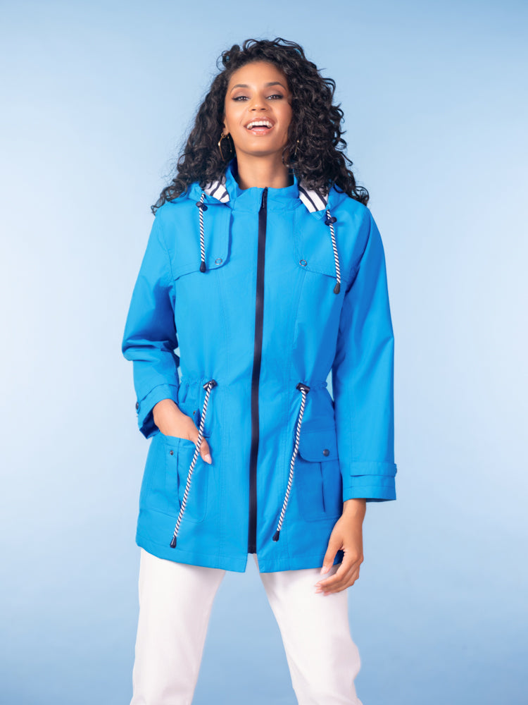 Women’s bright blue zip-up anorak jacket with hood and cinched waist detail, paired with light trousers against a studio backdrop.