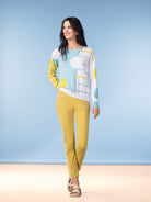 Model in a studio setting wearing a light blue and yellow abstract knit sweater with slim yellow pants and beige sandals.