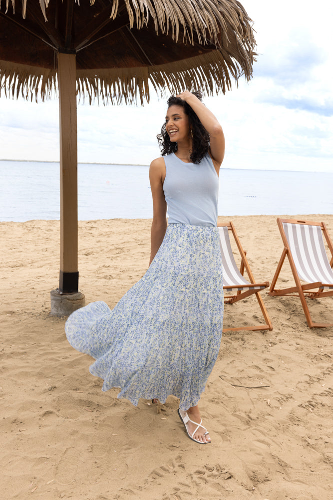 Model reclining outdoors in a light blue sleeveless knit tank with a soft drape and minimalist cut, styled with a blue floral skirt on a sandy beach.