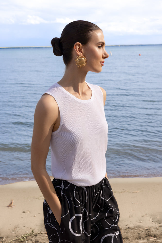 Woman standing on a beach wearing a white sleeveless knit top and black high-waisted pants with a white abstract print, gold sun-shaped statement earrings, and her dark hair styled in a sleek low bun, with the ocean in the background.