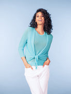 Woman wearing a teal draped cardigan-style top with white tailored trousers against a blue studio background.