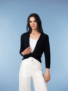 Studio image of a woman wearing a black lightweight open-front cardigan over a white tank top, paired with white jeans, photographed against a soft blue background.