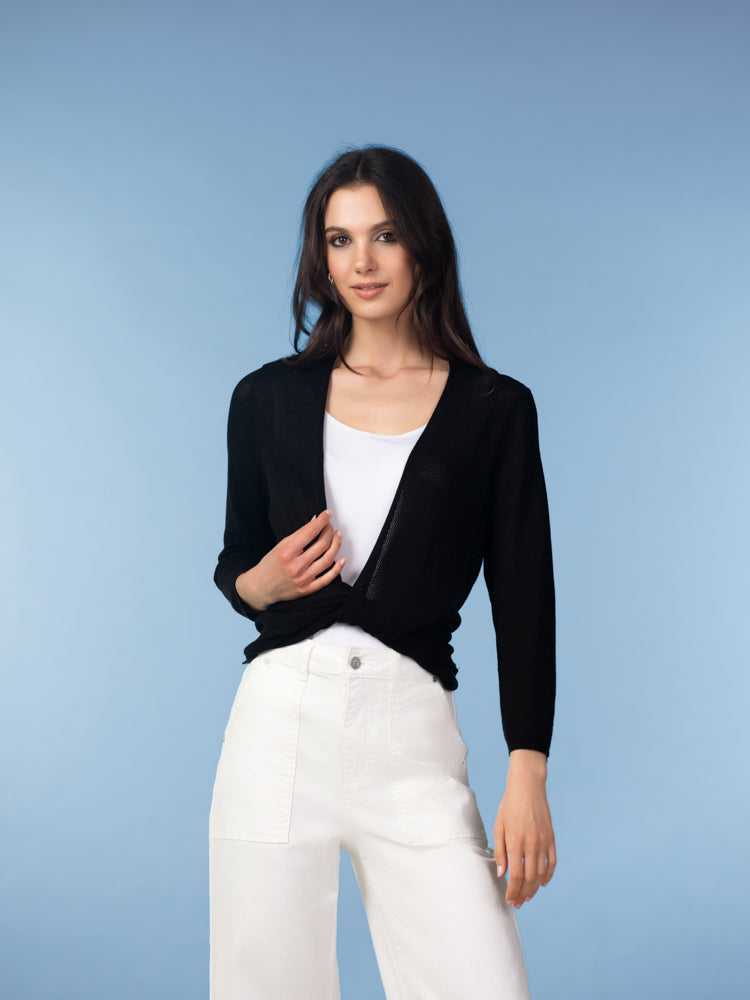 Studio image of a woman wearing a black lightweight open-front cardigan over a white tank top, paired with white jeans, photographed against a soft blue background.