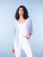 Woman wearing a light blue draped cardigan-style top with white tailored trousers against a blue studio background.