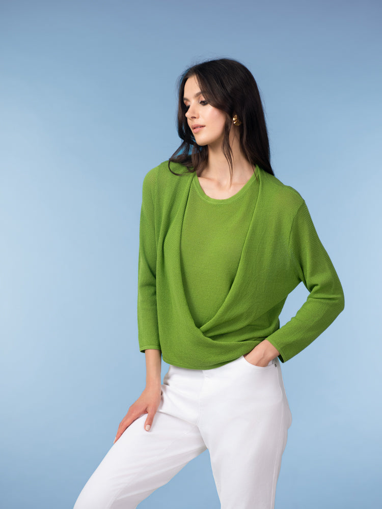 Woman wearing a green draped cardigan-style top with white tailored trousers against a blue studio background.