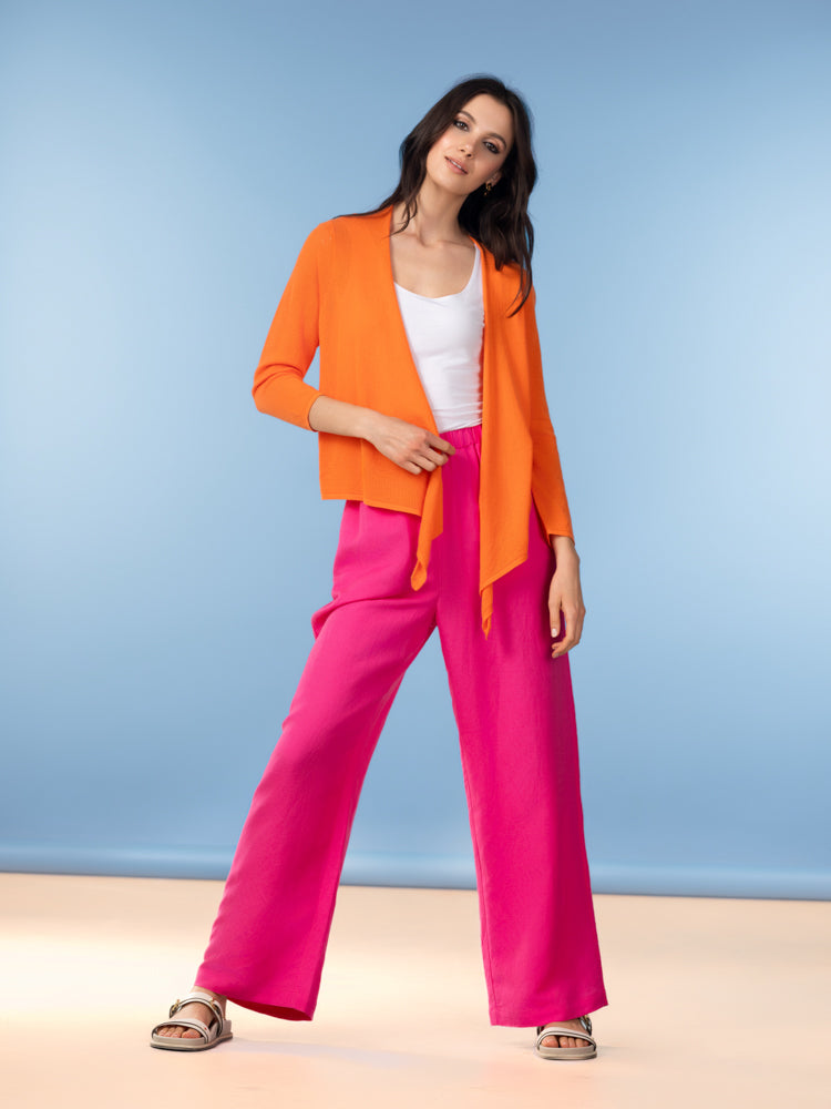 Woman wearing an orange lightweight cardigan over a white tank top with bright pink wide-leg trousers and beige sandals against a blue background.