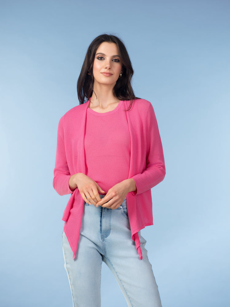 Woman wearing a pink open-front cardigan layered over a matching pink top with light blue jeans against a blue studio background.