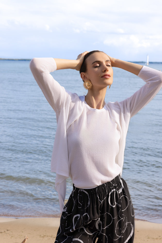 Woman wearing a white lightweight open-front cardigan with long sleeves, styled with black wide-leg cropped pants featuring a white abstract print, standing on a sandy beach with water in the background.