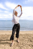 Full-length view of a woman in a white lightweight cardigan layered over a white top, paired with black cropped wide-leg pants with a white abstract pattern and neutral slide sandals, posing on a beach.