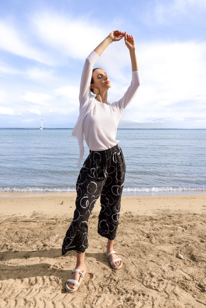 Full-length view of a woman in a white lightweight cardigan layered over a white top, paired with black cropped wide-leg pants with a white abstract pattern and neutral slide sandals, posing on a beach.