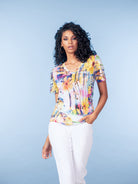 Woman modelling a short-sleeve V-neck T-shirt with a vibrant abstract floral print in yellow, pink, and blue tones, paired with light grey slim-leg trousers against a soft blue studio background.