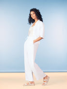 Model wearing a lightweight white linen blend button-front shirt with matching wide-leg pants and flat sandals, styled against a soft blue studio background.