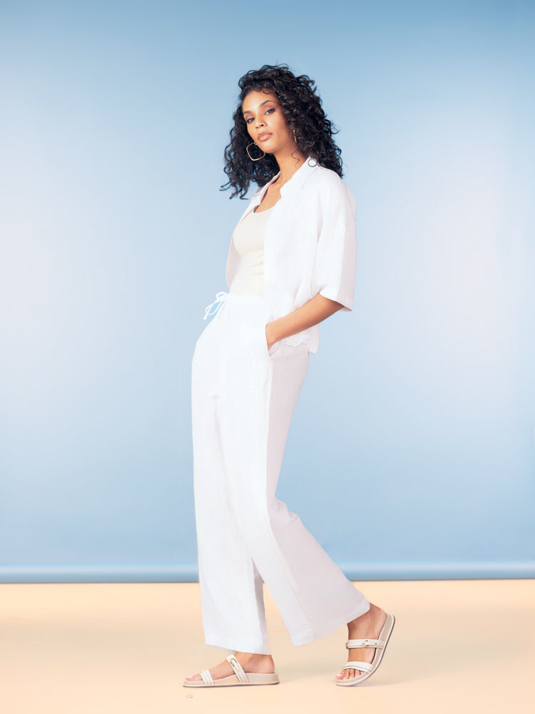 Model wearing a lightweight white linen blend button-front shirt with matching wide-leg pants and flat sandals, styled against a soft blue studio background.