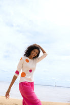 Model posing on the beach in a white knit sweater with orange and pink dot pattern and vibrant pink wide-leg trousers.