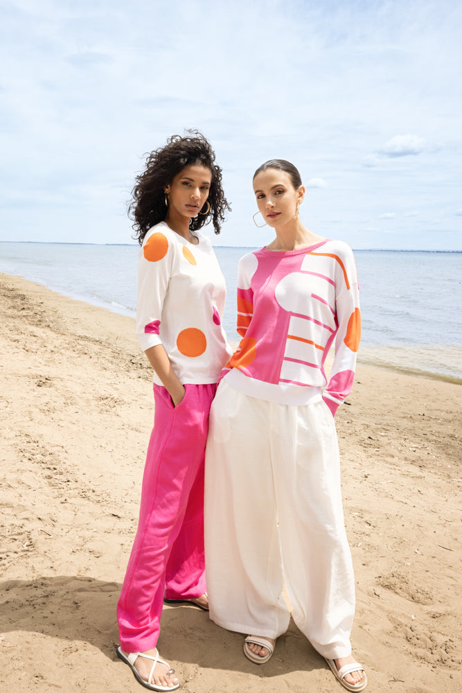 Two women standing on a beach wearing abstract knit sweaters; one styled in a white sweater with orange and pink dots and pink wide-leg pants, the other in a pink and white graphic sweater with white wide-leg trousers.