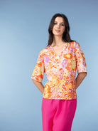 Woman modelling a vibrant abstract floral blouse in orange, pink, and soft lilac tones, featuring a V-neckline with delicate trim and elbow-length sleeves, paired with bright pink trousers against a light blue studio background.