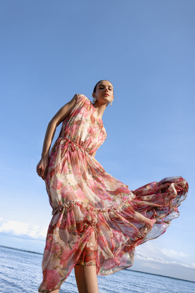 Low-angle view of a pink and coral floral print midi dress with flowing tiered hem, shown in motion on a beach.