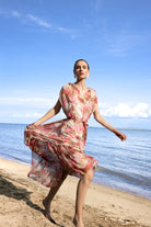 Model wearing a pink and coral floral print midi dress with tie waist, walking barefoot on a sandy beach.
