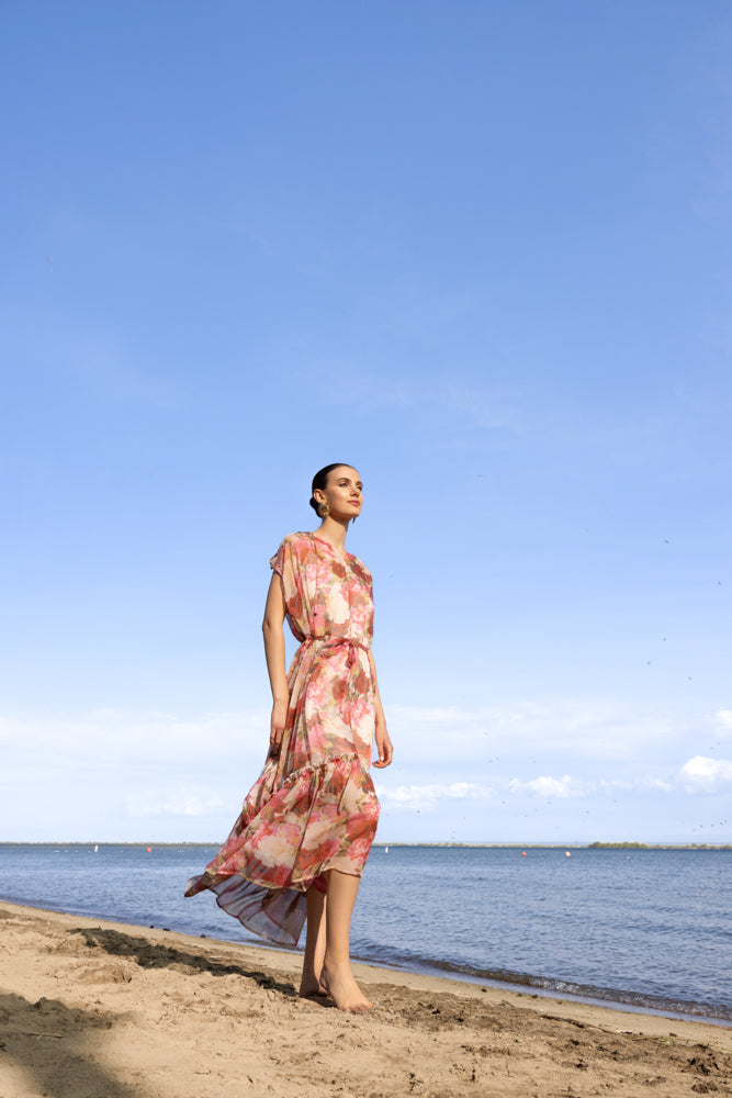 Model in a pink floral midi dress with cap sleeves and adjustable waist tie, standing by the water on a sunny day.