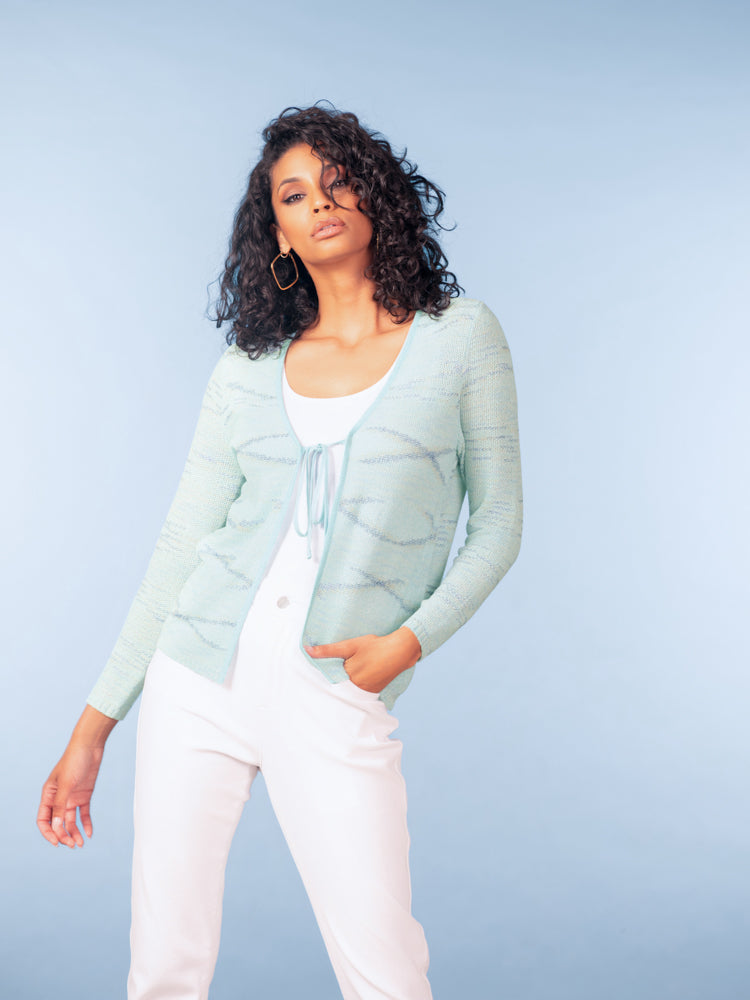 Woman wearing a light blue tie-front knit cardigan layered over a white top with white trousers, styled with gold hoop earrings against a soft blue studio background.