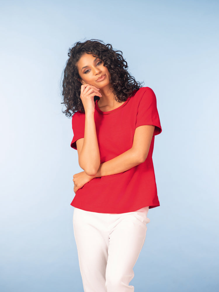 Model wearing a red short-sleeve cotton t-shirt with a round neckline and subtle trim detail, styled with light neutral trousers against a soft blue studio background.