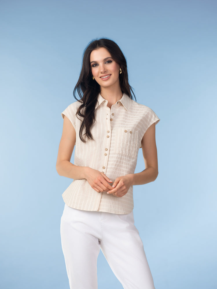 Woman wearing a beige textured cotton blouse with a collar and button-front closure, styled with light grey trousers against a soft blue background.