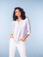 Model wearing a pastel striped three-quarter sleeve blouse in soft lavender, blue, and white tones, styled with white trousers against a light blue studio background.