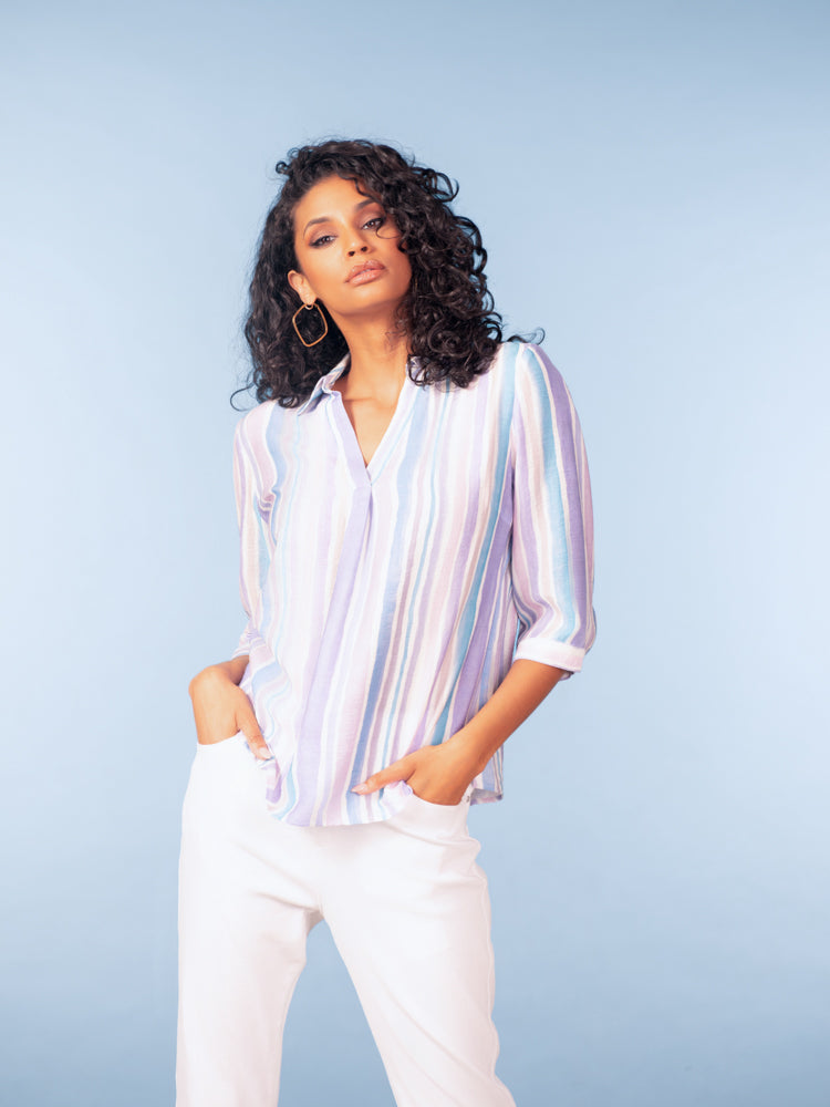 Model wearing a pastel striped three-quarter sleeve blouse in soft lavender, blue, and white tones, styled with white trousers against a light blue studio background.