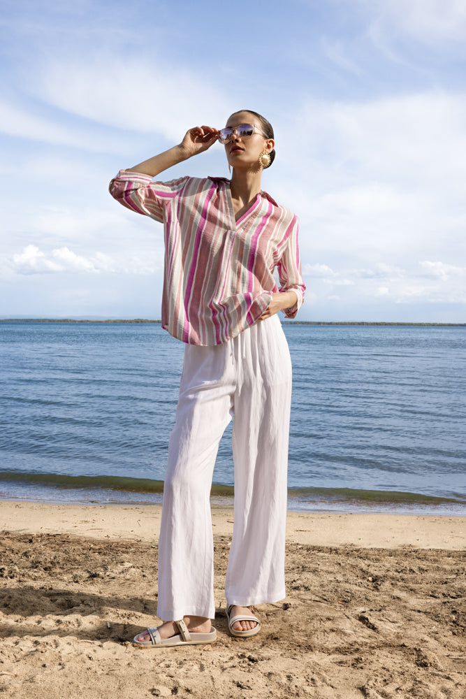 Model wearing a pink and white vertical striped three-quarter sleeve blouse with a relaxed fit, paired with wide-leg white trousers and sandals, standing on a sandy beach.