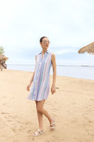 Model wearing a sleeveless striped button-front dress with a collared neckline and above-the-knee length, styled with clear-framed glasses and hoop earrings, shown from the back against a light sky background.