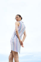 Model wearing a sleeveless striped button-front dress with a collared neckline and above-the-knee length, styled with clear-framed glasses and hoop earrings, shown from the back against a light sky background.