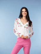 Woman wearing a white floral pointelle knit cardigan with a button-front V-neck and long sleeves, paired with pink pants, standing against a light blue background.
