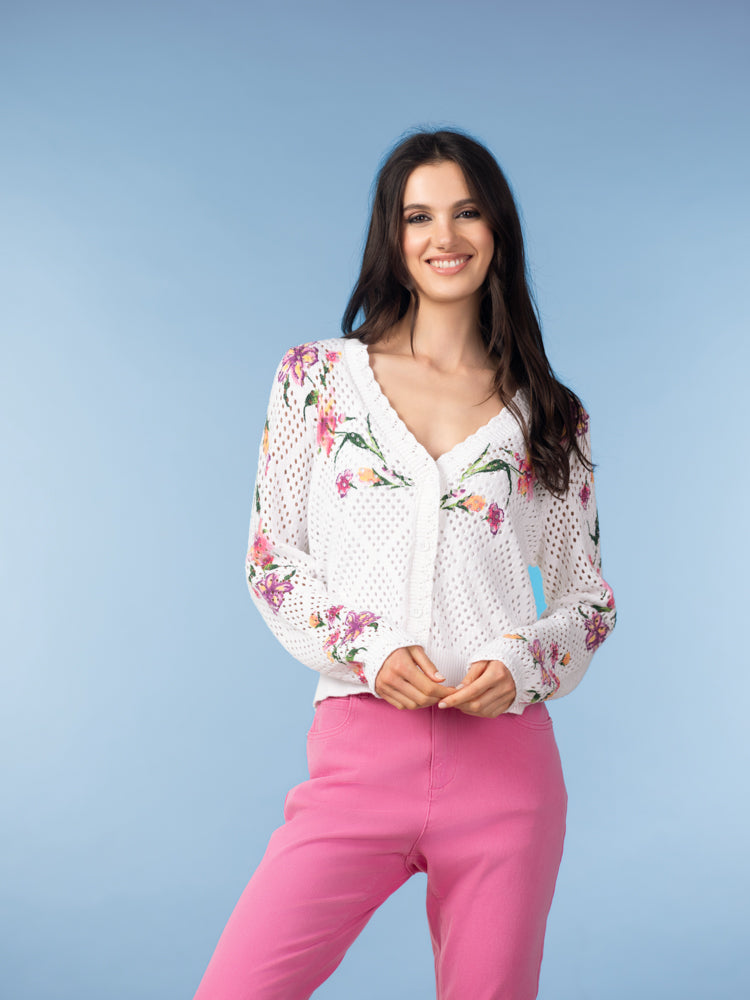 Woman wearing a white floral pointelle knit cardigan with a button-front V-neck and long sleeves, paired with pink pants, standing against a light blue background.