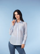 Woman modelling a lightweight button-front blouse with a classic collar and long sleeves, styled in a soft light blue tone. The blouse has a relaxed, fluid drape and is paired with fitted blue jeans against a matching studio backdrop.