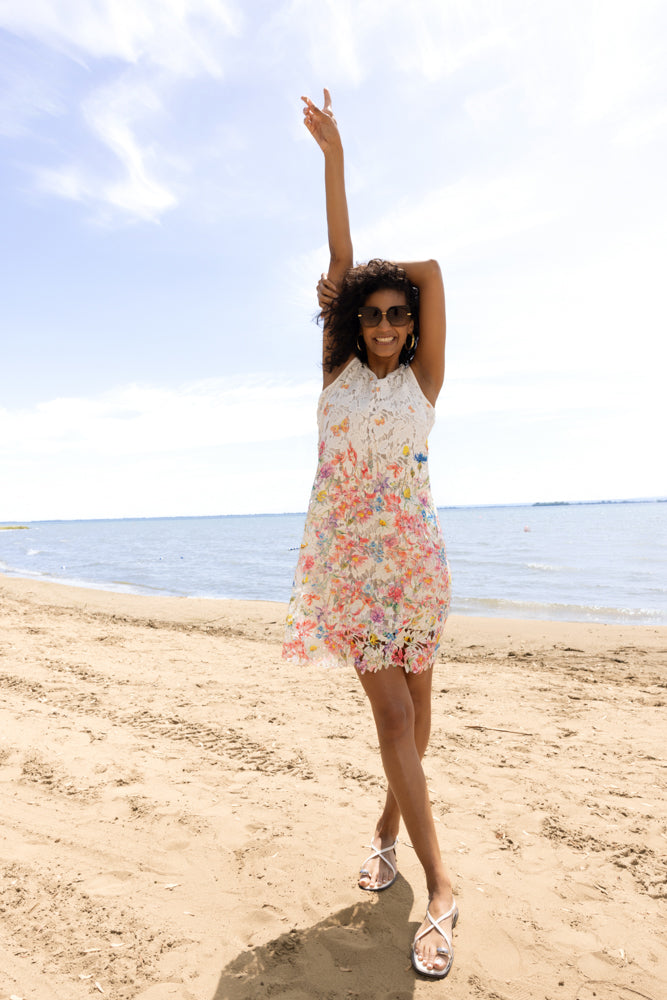 Woman wearing a sleeveless white floral lace shift dress with multicolour embroidery, posing on a sandy beach with the ocean in the background.